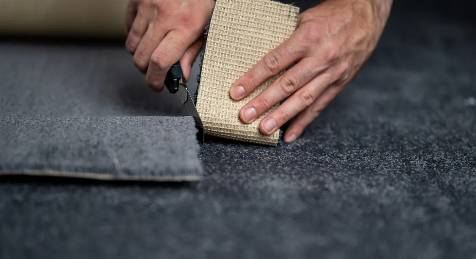 A carpet repair technician kneeling on the floor using a carpet knife to cut a clean section out of wall-to-wall carpet for a patch repair